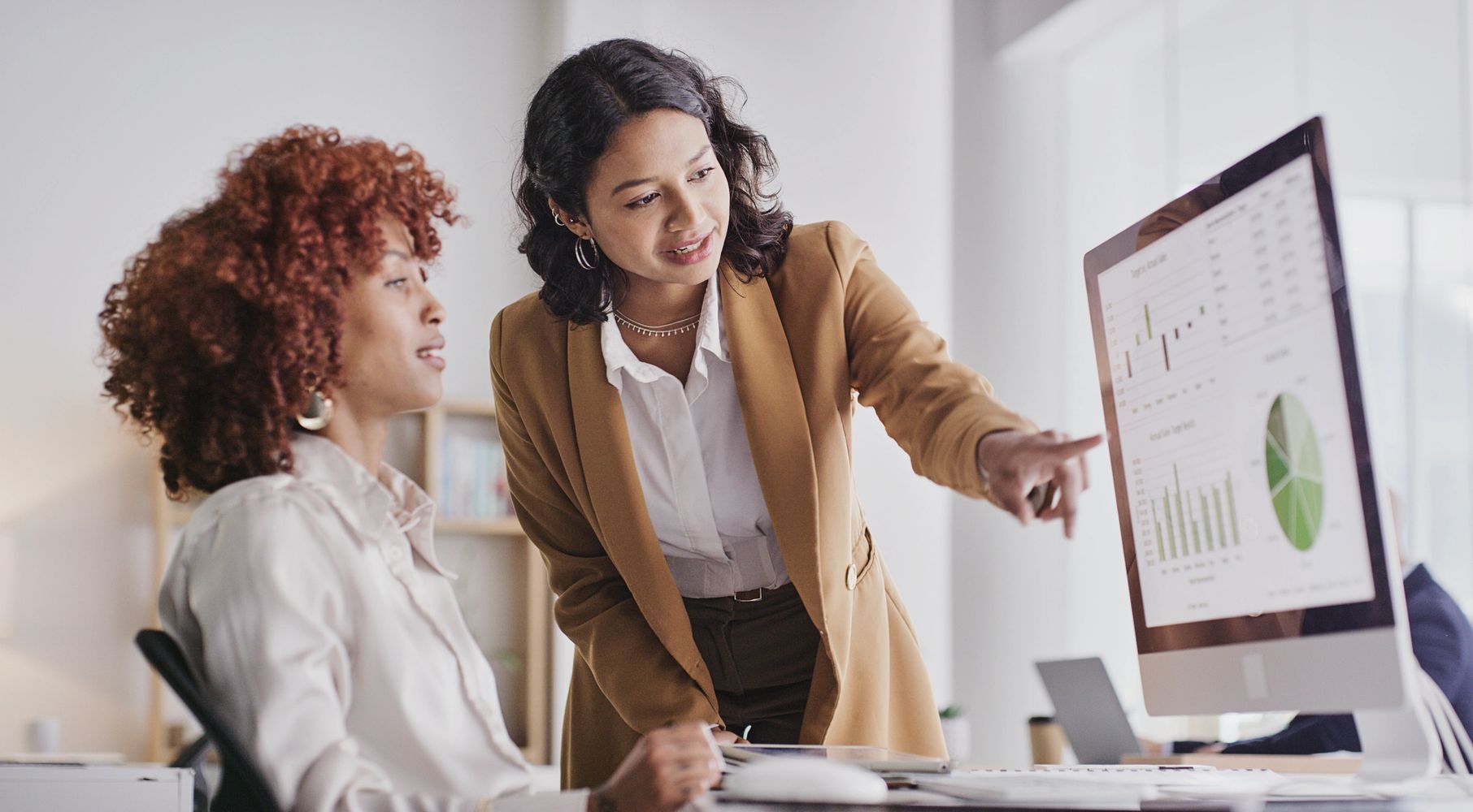 Two women analyzing data charts on a computer screen in an office.
