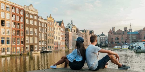 Two people sitting by a canal with historic buildings in the background.