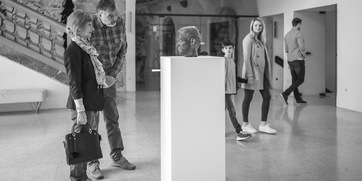 People observing a sculpture head displayed on a pedestal in a museum.