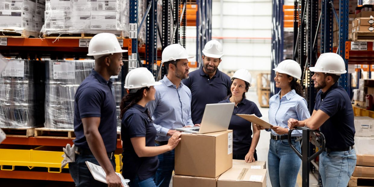 Warehouse team meeting with hard hats and laptops.