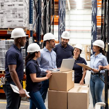 Team of warehouse workers discussing logistics with laptops and clipboards in storage area.