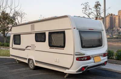A white camper trailer parked near a city riverbank with tall buildings in the background.