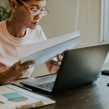A woman reviews documents while working on a laptop at a desk.