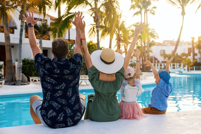 happy family with kids and arms raised having fun by the swimming pool at tropical resort hotel. summer vacation. rear view