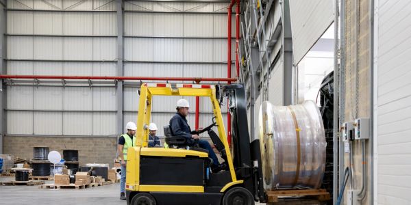 Forklift lifting large metal spool in warehouse.