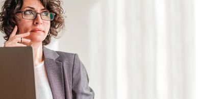 Woman in glasses working thoughtfully at a laptop with pen and paper.