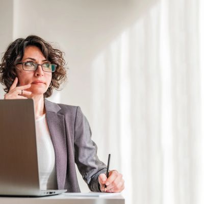 Thoughtful woman in glasses working on a laptop in a bright office.