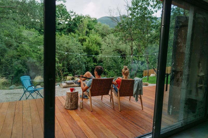 Photo of a heterosexual couple resting and working on a deck in front of their cabin; Young woman is working remotely on her computer while her partner is playing guitar