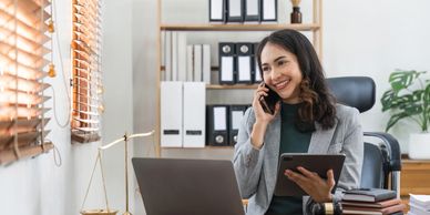 A woman in business attire talking on phone while holding a tablet in an office.