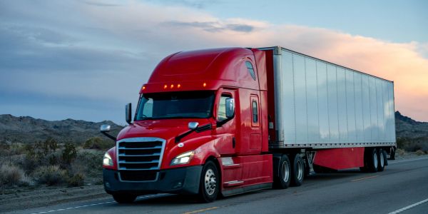 Red semi-truck driving on a desert highway at sunset.
