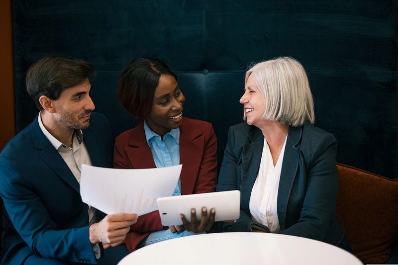 Positive diverse colleagues in formal suits sitting at white round table with document and tablet and discussing business strategy in office cafeteria