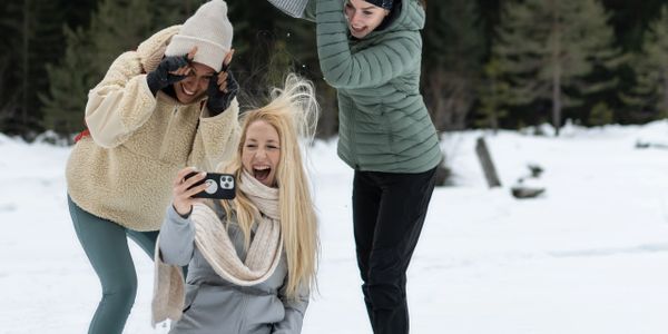 Three friends having fun in the snow, one taking a selfie while the others playfully tease her.