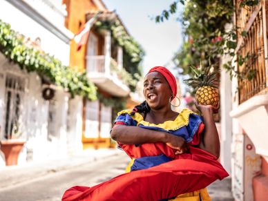 Mujer afroamericana con vestido tradicional y piña en la mano sonríe en la calle.