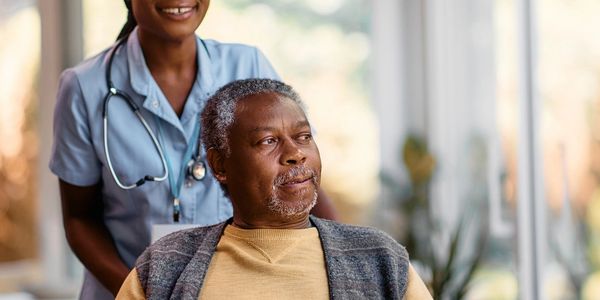 Nurse smiling behind elderly man in wheelchair indoors.