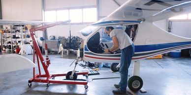 Man repairing a small airplane engine in a hangar using a red hoist.