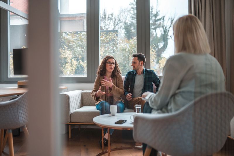 Candid shot through glass, Caucasian couple explaining something to a blond female professional, only her back visible through glass of the office.