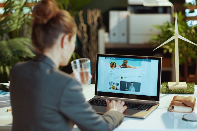Seen from behind modern woman with laptop in modern office reading news on social media.