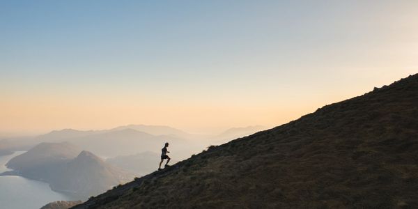 A lone runner ascends a steep hill at sunrise with mountains and a lake in the background.