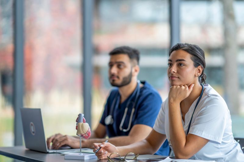 A small group of medical students are seen sitting at desks in a classroom during a lesson.  They are each wearing scrubs and paying close attention.