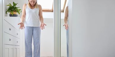 A woman in pajamas looks disappointed standing on a scale in a bright room.