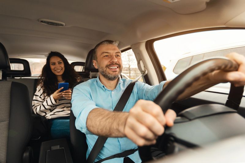 Portrait of happy male driver riding car, cheerful beautiful lady sitting inside auto on back passenger seat, using cell phone and looking on road talking with guy, enjoying safe ride, windshield view
