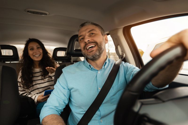 Nice Ride. Portrait of happy male driver riding car looking at cheerful beautiful lady sitting inside auto on back passenger seat, female using cell phone and talking with guy, windshield view
