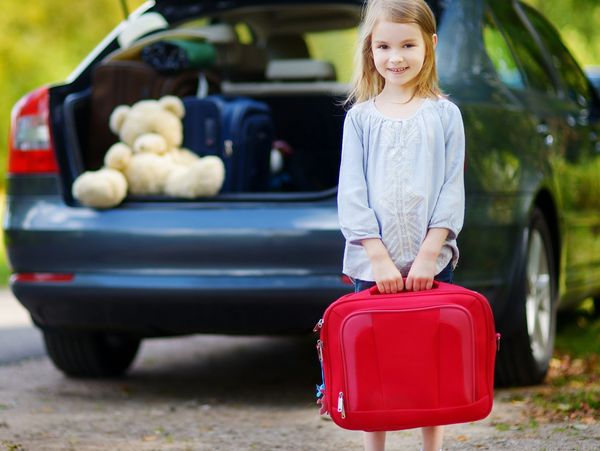 Smiling girl holding red suitcase in front of open car trunk packed for travel.