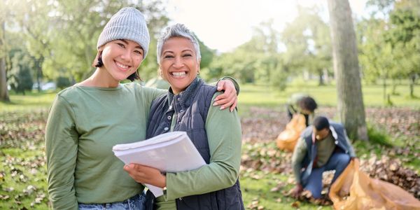 Two women smiling outdoors, one holding papers, with people cleaning leaves in the background.