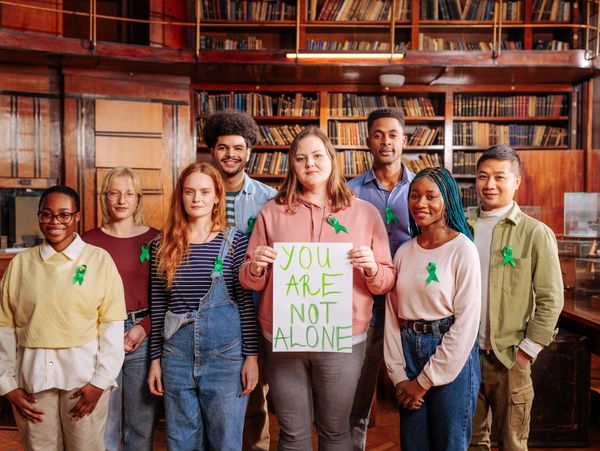 Diverse group wearing green ribbons holds a sign saying 'You Are Not Alone' in a library.