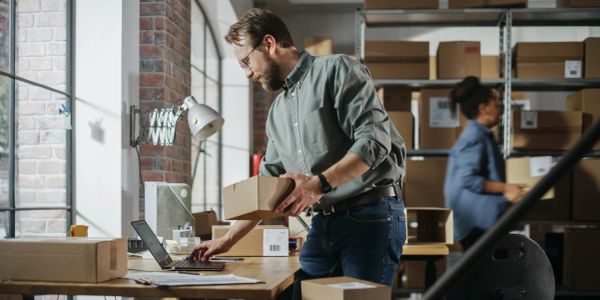 Man packing boxes while working on a laptop in a warehouse.