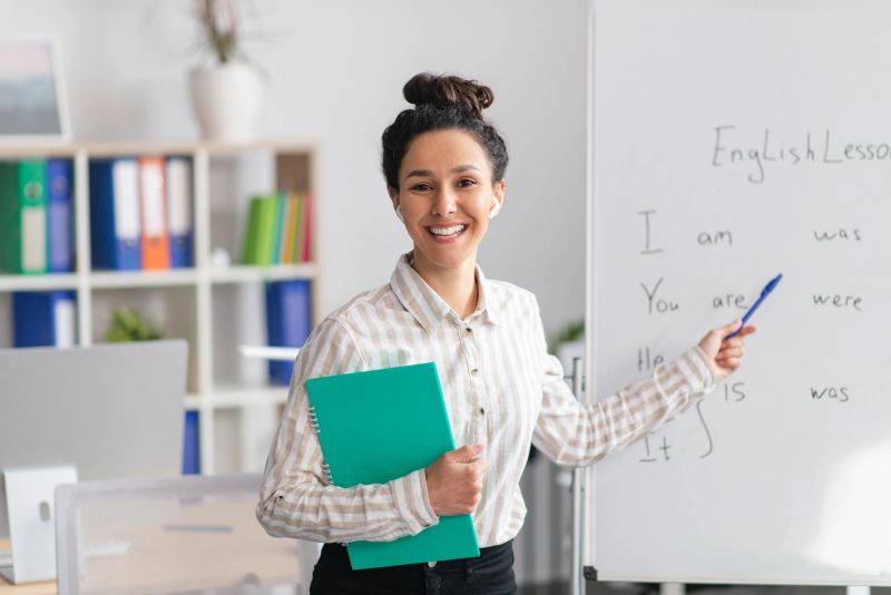 Training concept. Happy female tutor teaching English language, pointing at grammar rules on board and smiling at camera, holding clipboard in office interior