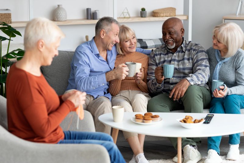 Positive multiracial senior people men and women spending time together at board and care home, drinking tea, eating cookies, laughing and having conversation, knitting, enjoying free time