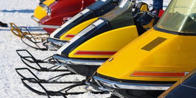 Lineup of colorful vintage snowmobiles on snow with people in the background.