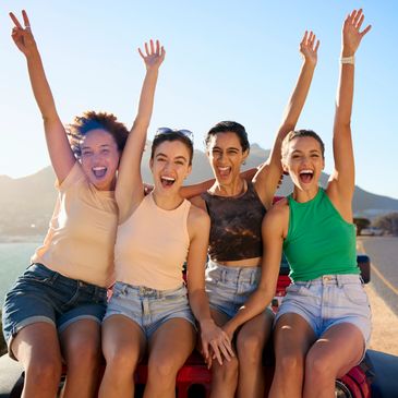 Four friends joyfully raising their hands while sitting outdoors on a sunny day.