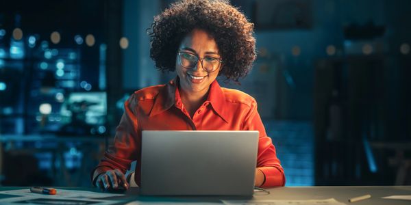 A woman in an orange shirt smiles while working on a laptop at night.