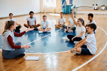 A teacher and students sitting in a gym circle, girl raising her hand.