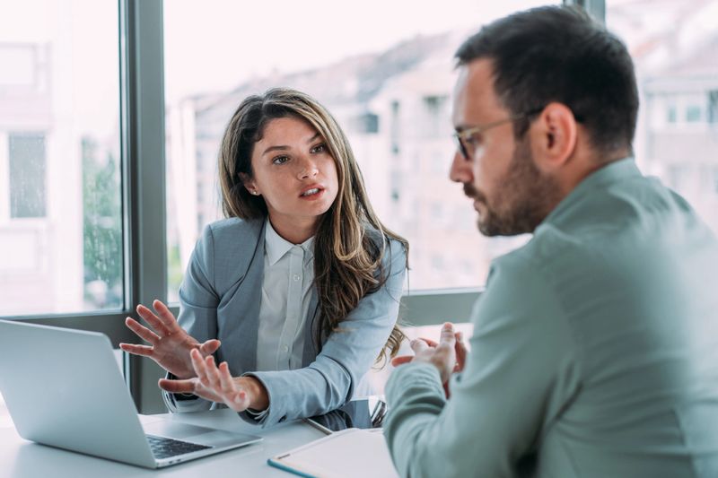Shot of a two confident business persons sitting on a desk in the office and sharing ideas. Businessman and businesswoman in meeting using laptop and discussing business strategy. Business coworkers working together in the office.