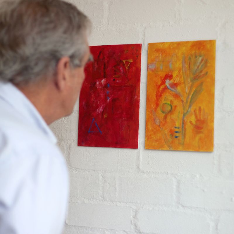 Square shot of an adult male looking closely at a red and a yellow abstract painting on a white wall.