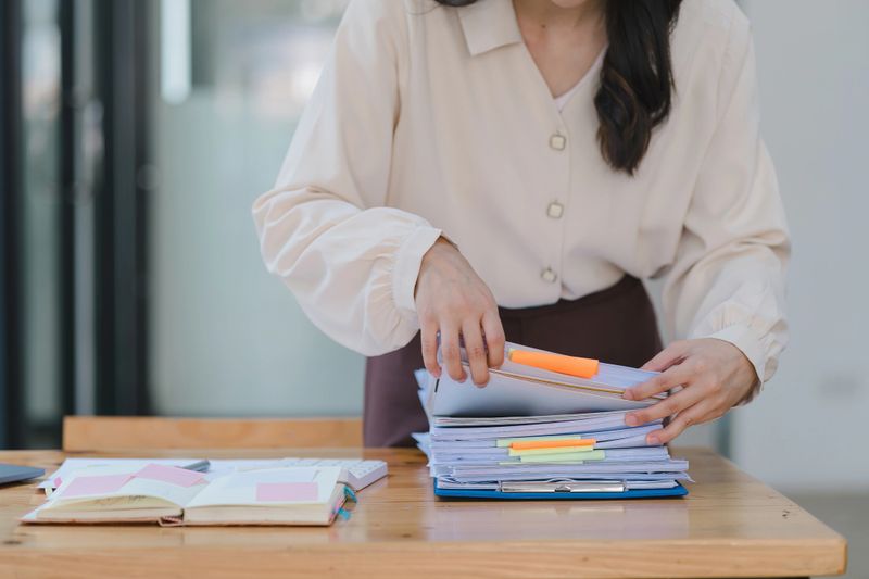 A businesswoman is sifting through stacks of paper files and folders that contain both incomplete and completed documents