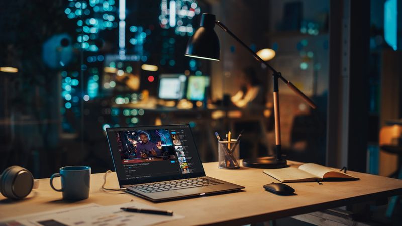 Laptop Standing on a Desk with a Video Streaming Platform Displayed on Screen. Table with Computer, Coffee Mug, Headphones and Notebook in Creative Office at Night.