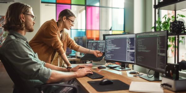 Two colleagues collaborating on coding at a dual-monitor workspace in a colorful office.