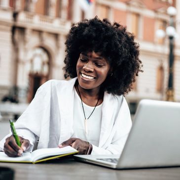 Gen AI image of a smiling black woman writing in her notebook with a laptop in front of her outside.