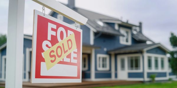 For sale sign with additional sold sign placed on top, in front of blue house.