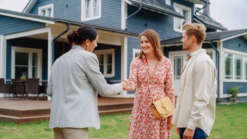 Happy Couple Shaking Hands with real estate agent in Front of Their New Home. Professional Real Estate Agent Congratulating Young Homeowners with Property Purchase, Giving Them the Key to the House.