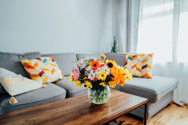 Vase with huge multicolor various flower bouquet on the coffee table with blurred background of modern cozy light living room with gray couch sofa. Giving flowers. Mother's day, birthday gift