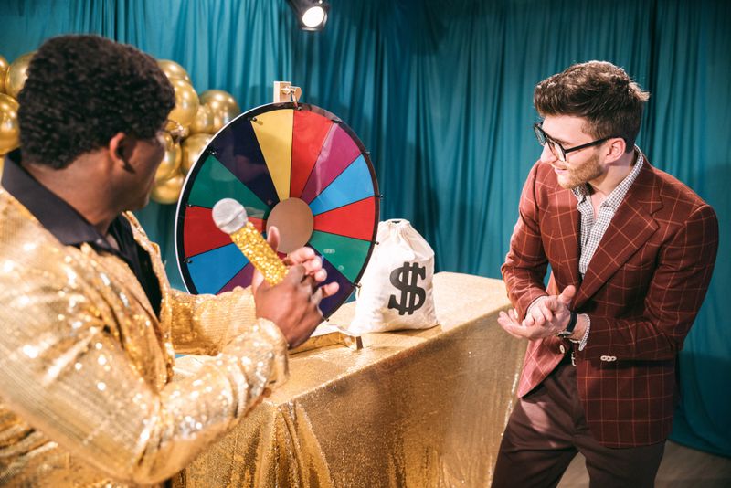A fun depiction of a competitive TV game show, stylized in late 1970's or early 1980's fashion.  The host, an African American man in a stunning gold blazer, watches as contestants spin a colored wheel to win cash prizes.