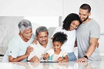 Smiling multi-generational family gathered around a tablet at the kitchen counter.
