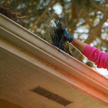 Person cleaning debris from a house gutter wearing a blue patterned glove and pink sleeve.