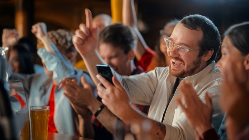 Excited Man in Glasses Sitting at a Bar Stand, Using a Smartphone, Feeling Alive After Putting a Bet on His Favorite Soccer Team. Ecstatic When Football Team Scores a Goal and He Wins the Prize.