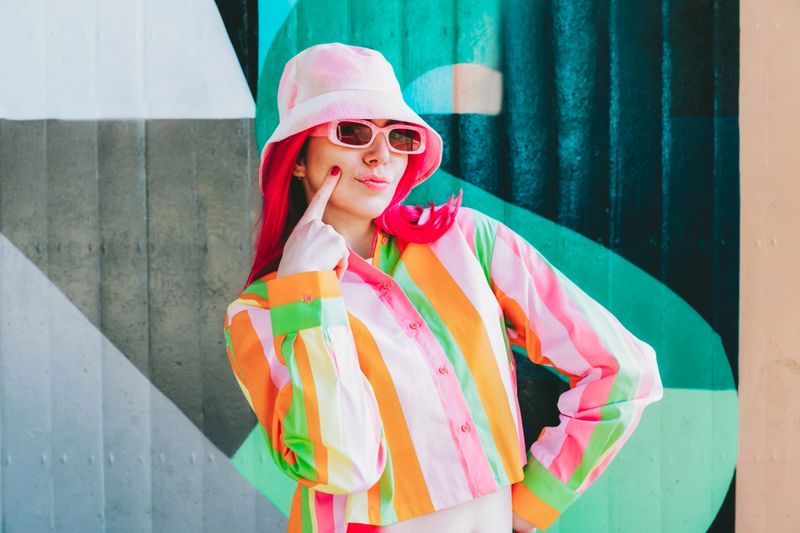 Urban Fresh street fashion look. Vanilla Girl. Kawaii vibes. Candy colors design. Bucket hat trends. Young woman with pink hair and sun glasses in multicolor strippled shirt on the wall background.